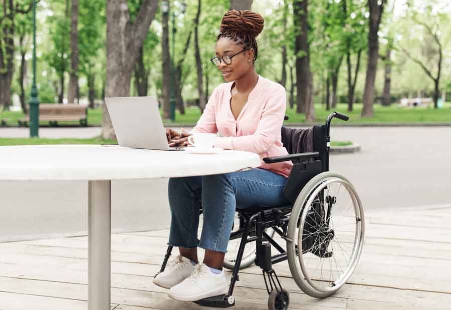 a woman in a wheelchair sits at a table in an open air cafe using a laptop.